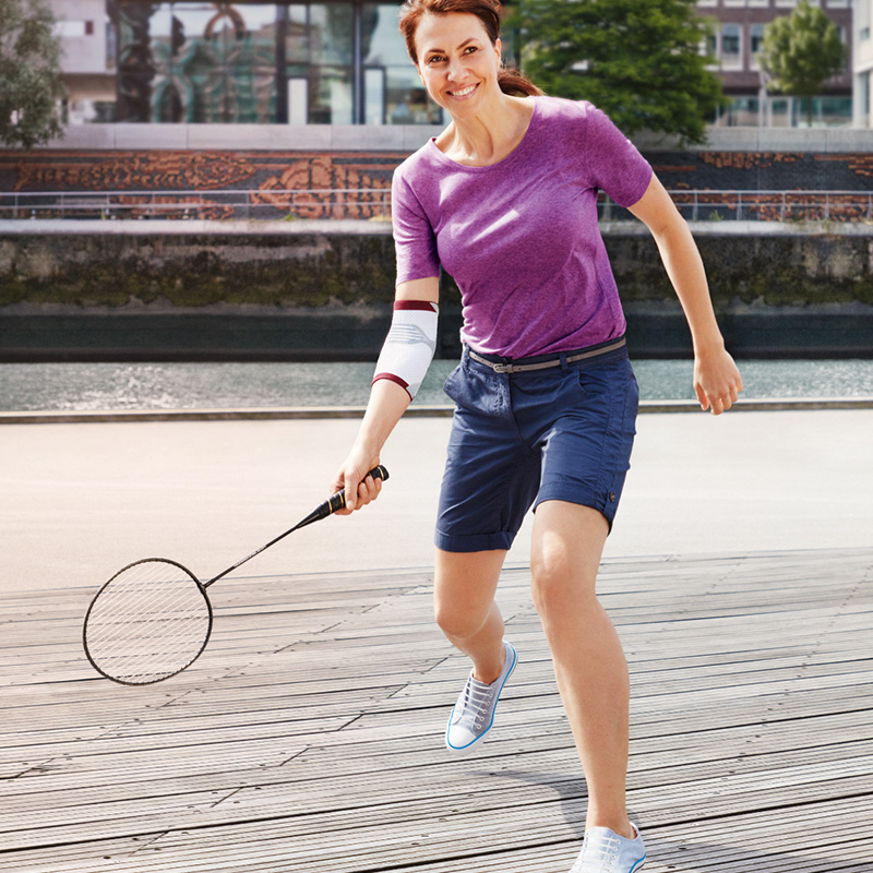 Woman wearing an elbow support and purple top and blue shorts prepares to hit a ball with her racket